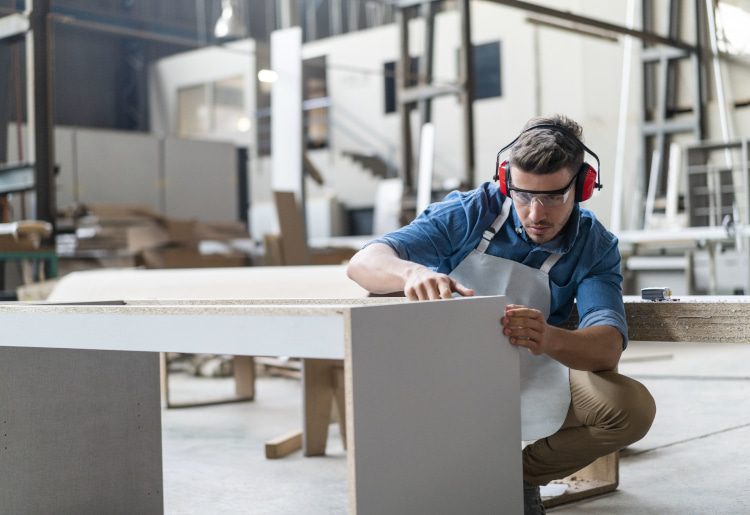 Skilled carpenter making closet in carpentry workshop. Young craftsman is examining wood. He is working in factory.